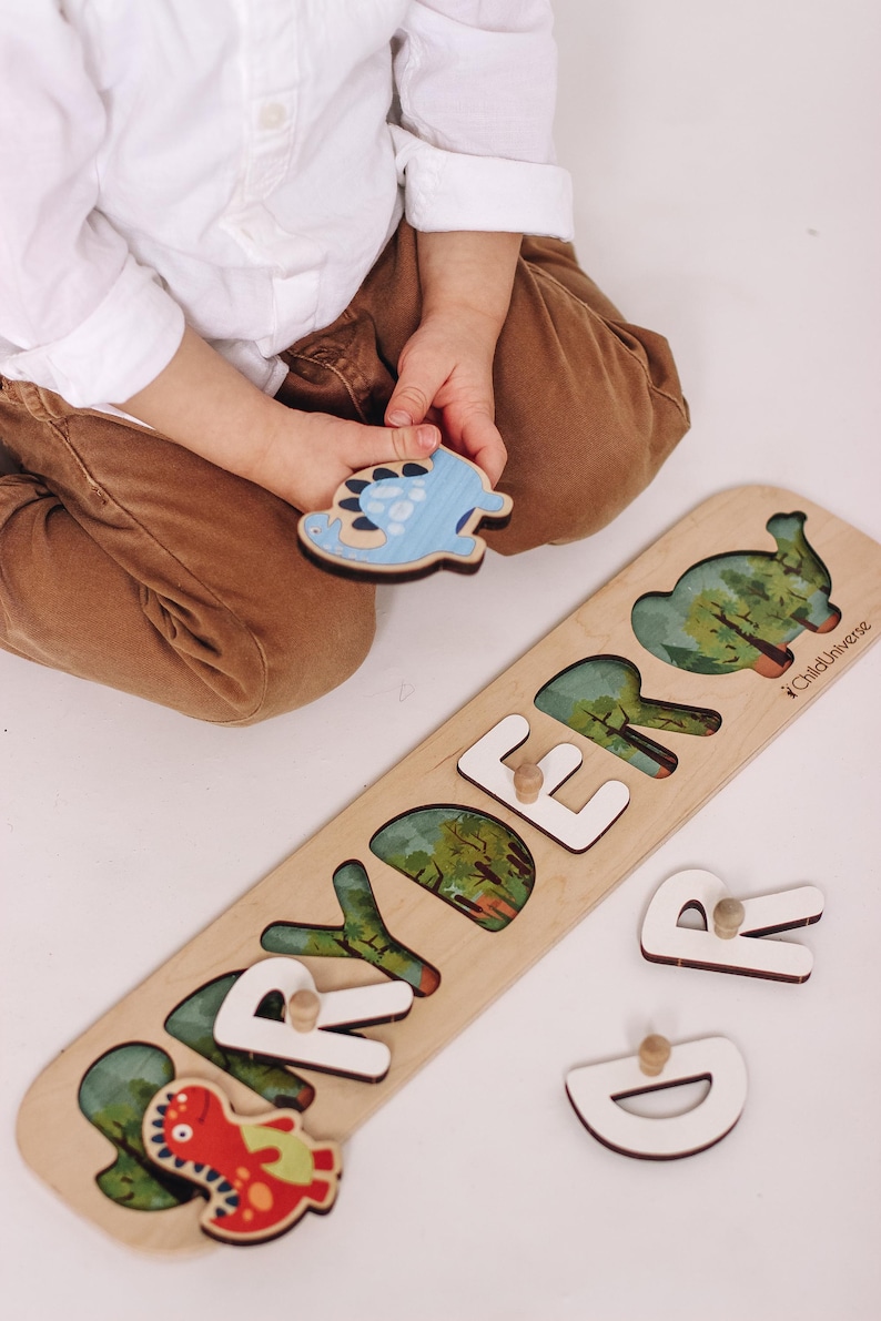 May include: Wooden dinosaur puzzle with the name "RYDER" spelled out in white letters. The puzzle pieces feature colorful dinosaur illustrations and a light wood finish. A child is holding a blue dinosaur piece.
