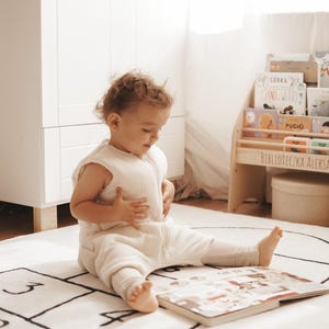 May include: A cream-colored sleeveless wearable blanket with a front zipper closure, worn by a toddler sitting on a white rug featuring a black hopscotch grid pattern while looking at an open book.