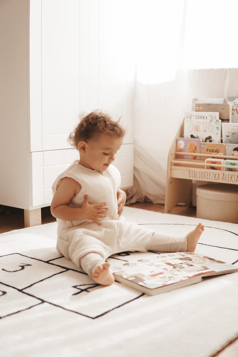 May include: A cream-colored sleeveless wearable blanket with a front zipper closure, worn by a toddler sitting on a white rug featuring a black hopscotch grid pattern while looking at an open book.