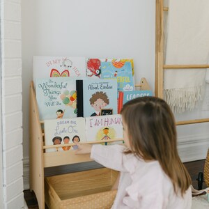 May include: A light-colored wooden bookshelf with two tiers and a storage bin at the base. The shelves hold several children's books with titles including The Wonderful Things You Will Be, Albert Einstein, Dream Big, Little One, and I Am Human.