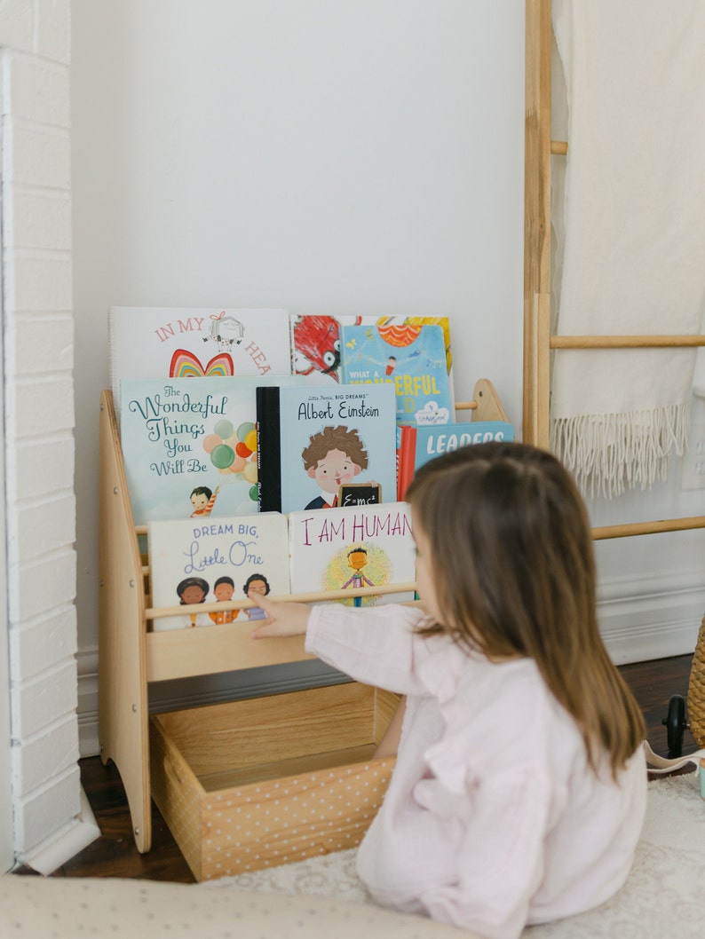May include: A light-colored wooden bookshelf with two tiers and a storage bin at the base. The shelves hold several children's books with titles including The Wonderful Things You Will Be, Albert Einstein, Dream Big, Little One, and I Am Human.