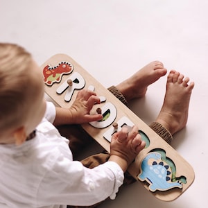 May include: Wooden name puzzle with the name "GRACE" spelled out in white letters. The puzzle features a red dinosaur, a blue dinosaur, and wooden pegs for easy handling. A fun and educational toy for toddlers.