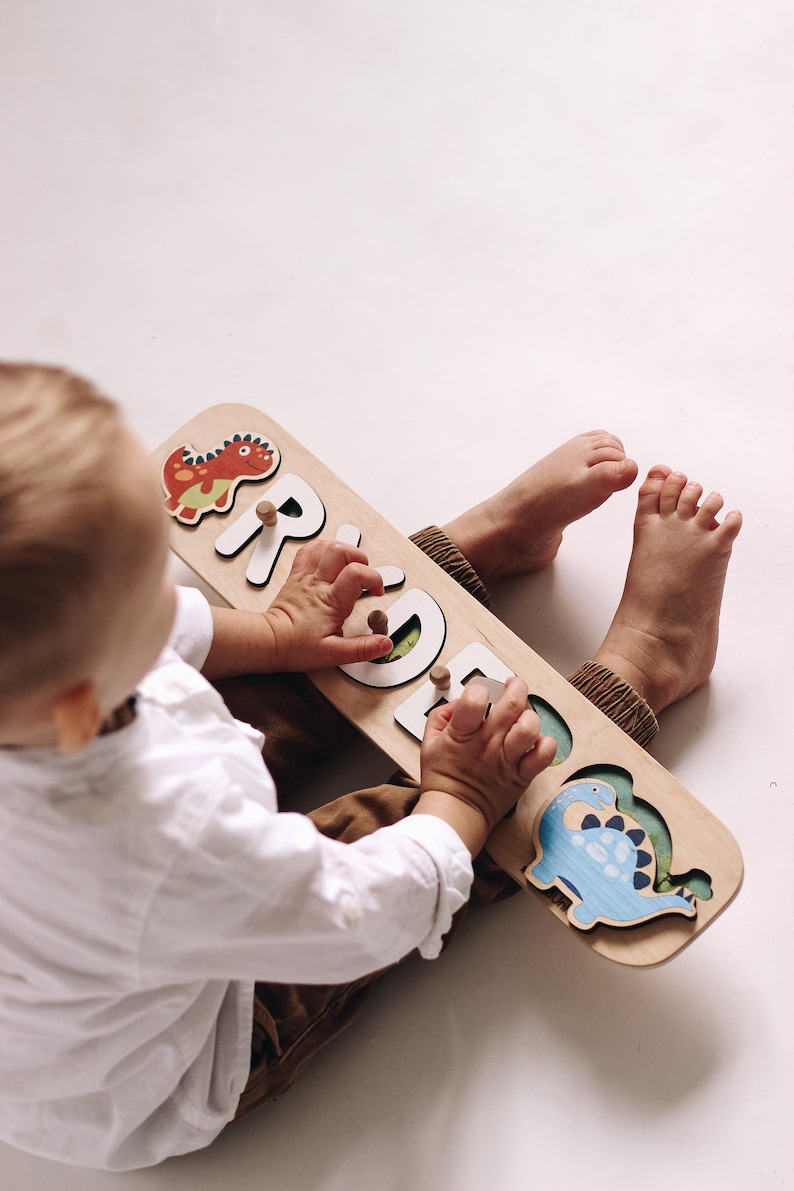 May include: Wooden name puzzle with the name "GRACE" spelled out in white letters. The puzzle features a red dinosaur, a blue dinosaur, and wooden pegs for easy handling. A fun and educational toy for toddlers.