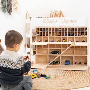 K&ouml;nnte beinhalten: Ein Holzspielzeug-Garage mit mehreren F&auml;chern, gef&uuml;llt mit Miniatur-Spielzeugautos. Die Garage ist mit "Oliver's Garage" beschriftet. Ein Kind sitzt vor der Garage und spielt mit Spielzeugautos auf einer gewebten Matte.