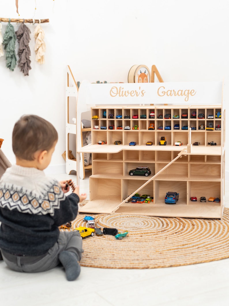 K&ouml;nnte beinhalten: Ein Holzspielzeug-Garage mit mehreren F&auml;chern, gef&uuml;llt mit Miniatur-Spielzeugautos. Die Garage ist mit "Oliver's Garage" beschriftet. Ein Kind sitzt vor der Garage und spielt mit Spielzeugautos auf einer gewebten Matte.