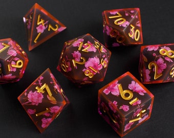 May include: Set of seven pink and brown dice with gold numbers and floral designs. The dice are a d4, d6, d8, d10, d%, d12, and d20.