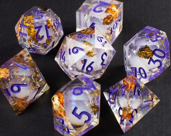 May include: Set of seven iridescent dice with gold flecks and purple numbers. The dice are clear and show a white interior. The dice are on a black surface.