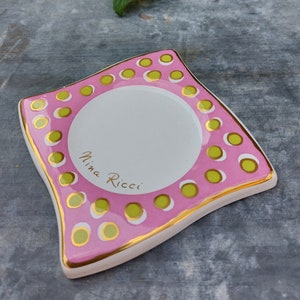 Peut inclure: Un plat en céramique rose et blanc avec un bord doré et des pois verts. Le plat a un centre surélevé et les mots "Nina Ricci" sont écrits en lettres dorées.