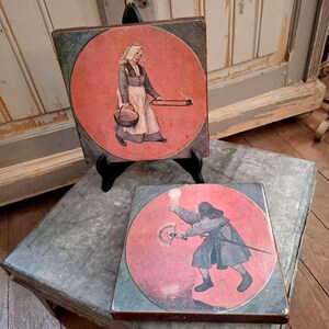 May include: Two vintage wooden art pieces. Each features a circular painting on a square base. One depicts a figure carrying a basket and a smoking object. The other shows a figure with a fan and a sword, set against a red backdrop.