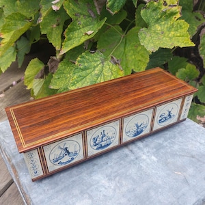 May include: A wooden box with a dark brown finish and a gold trim. The box has five decorative tiles with blue and white Delftware designs. The tiles depict windmills, houses, and other Dutch scenes.