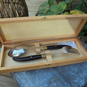 Peut inclure: Une boîte en bois avec un couvercle à charnière, contenant un service de salade en argent avec des manches noirs. La boîte est ouverte et le service est visible à l'intérieur. La boîte porte une petite étiquette avec le texte "Les Métiers d'Art en Layon".