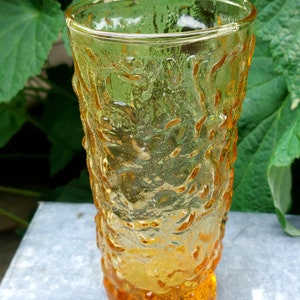 Könnte beinhalten: Ein gelbes Glas aus Glas mit einer strukturierten Oberfläche. Das Glas hat einen abgerundeten Boden und einen geraden, zylindrischen oberen Teil.
