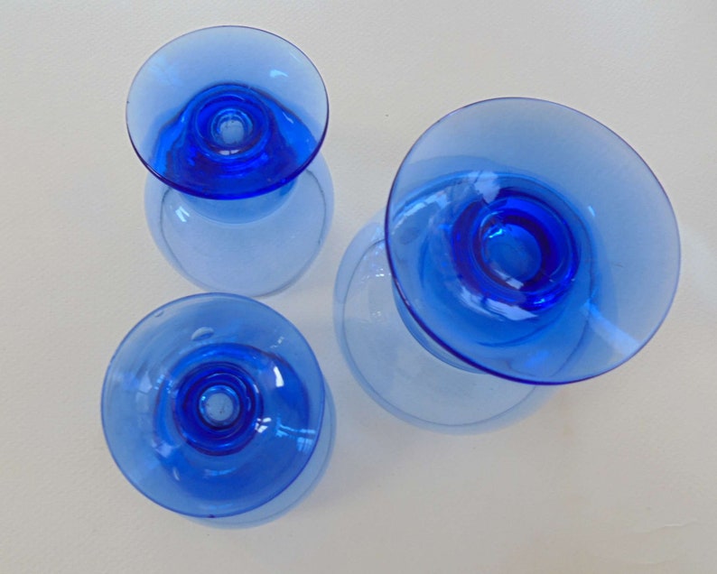 Op de afbeelding: Drie blauwe glazen kaarshouders met een brede basis en een smalle bovenkant. De kaarshouders zijn gemaakt van helder glas met een blauwe tint.
