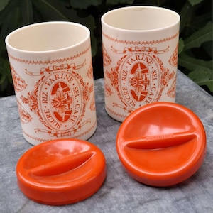Puede incluir: Dos botes blancos vintage con letras y diseños naranjas, incluyendo las palabras "SUCRE, RIZ, FARINE, SEL, CAFE". Cada bote tiene una tapa naranja con un asa. Los botes son cilíndricos.