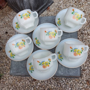 May include: Six white teacups with floral designs in shades of yellow, blue, and orange. Each cup is on a matching saucer with a gold rim.