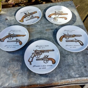Puede incluir: Cinco platos de cerámica blanca con ilustraciones en negro y marrón de pistolas y revólveres antiguos. Cada plato presenta un tipo de arma de fuego diferente con texto en francés que describe el tipo de pistola y el siglo en que se fabricó.