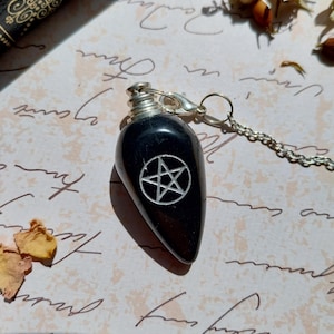 May include: A black pendulum with a silver chain and cap. The pendulum has a teardrop shape and features a white pentagram symbol. The background includes dried flowers and handwritten script on a light-colored surface.