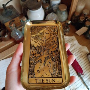 Pu&ograve; includere: Un vassoio rettangolare dorato con un disegno di carta dei tarocchi. La carta presenta un sole con un volto, un cavallo e il testo "THE SUN". Il vassoio viene probabilmente utilizzato per contenere piccoli oggetti.