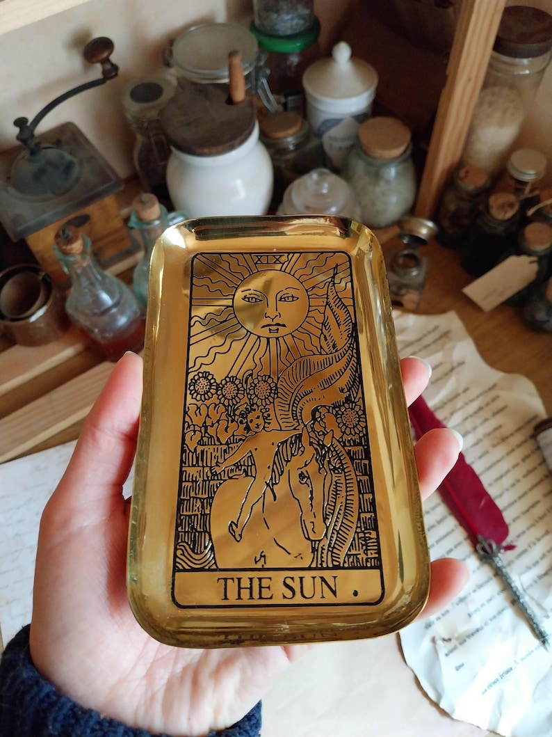 Pu&ograve; includere: Un vassoio rettangolare dorato con un disegno di carta dei tarocchi. La carta presenta un sole con un volto, un cavallo e il testo "THE SUN". Il vassoio viene probabilmente utilizzato per contenere piccoli oggetti.