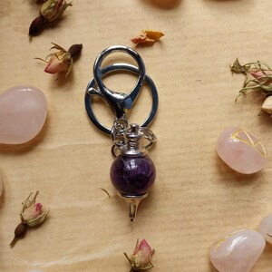 Puede incluir: Un péndulo plateado con una esfera de amatista de color púrpura intenso y una punta. El péndulo está rodeado de piedras de cuarzo rosa y capullos de rosa secos sobre una superficie beige.