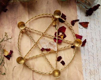Puede incluir: Un portavelas de pentagrama de metal dorado con cinco copas circulares para velas. Un cristal transparente y pétalos de rosa secos están dentro del pentagrama. El soporte se encuentra sobre una superficie similar al pergamino, con frascos y hierbas secas en el fondo.