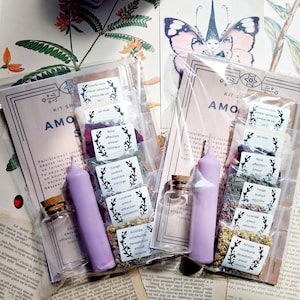 Könnte beinhalten: Ein Set für Liebeszauber mit einer rosa Kerze, einer kleinen Glasflasche und verschiedenen Kräutern und Kristallen. Das Set enthält Rosenblätter, Lavendel, Salbei und Schafgarbe. Das Set ist mit "AMO KIT SPELLS" beschriftet und enthält Anweisungen auf Französisch.