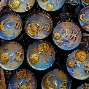Puede incluir: Una colección de velas redondas de lata negra con cera azul y dorada. Cada vela presenta un símbolo del zodiaco dorado diferente, un sol, una luna y estrellas. Las velas están dispuestas sobre una superficie oscura, creando un tema celestial.