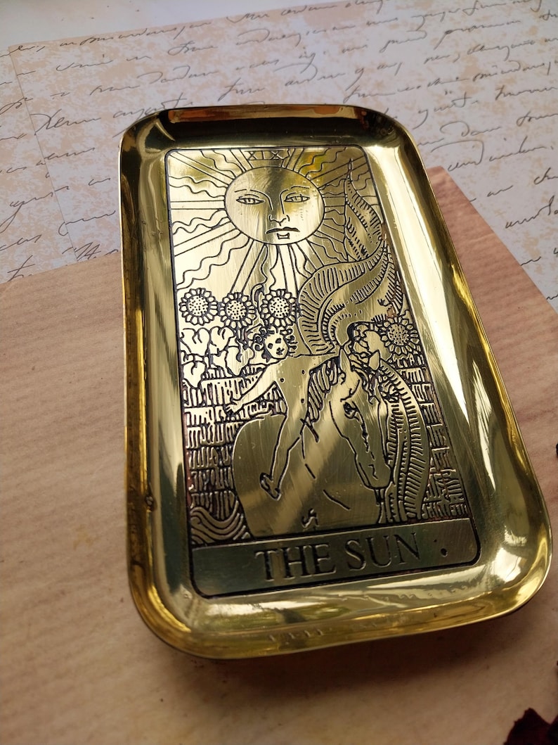 Pu&ograve; includere: Un vassoio rettangolare in ottone con angoli arrotondati. Il vassoio presenta un disegno inciso del sole con un volto, fiori e una persona a cavallo. Le parole "THE SUN" sono nella parte inferiore del vassoio.