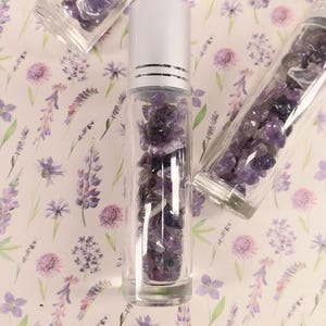 Peut inclure: Un flacon roll-on en verre transparent rempli de pierres d'améthyste violettes, surmonté d'un bouchon argenté. Deux autres tubes en verre remplis d'améthyste sont également présents. Le fond est un motif floral avec des fleurs lavande et violettes.