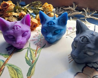 Puede incluir: Tres velas con forma de gato con un tercer ojo en la frente. Las velas son moradas, azules y negras.