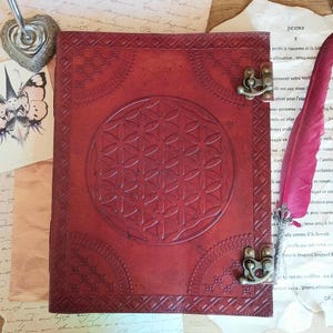 Puede incluir: Un diario de cuero rojo con un diseño geométrico de la flor de la vida en relieve en la portada. El diario tiene patrones decorativos en las esquinas, cierres metálicos y una pluma de pluma. El fondo incluye papeles vintage.