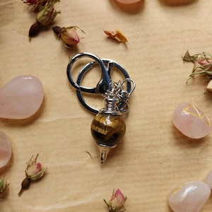 May include: A pendulum with a silver metal chain and a tiger's eye stone sphere. The pendulum is surrounded by pink rose quartz stones and dried rose buds on a tan surface.