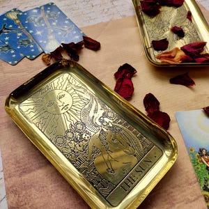 Pu&ograve; includere: Vassoio rettangolare color oro con un'immagine incisa del sole e le parole "THE SUN". Il vassoio &egrave; circondato da petali di rosa secchi e carte dei tarocchi. Il vassoio viene probabilmente utilizzato per scopi decorativi o spirituali.