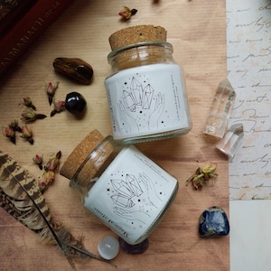 Pode incluir: Dois frascos de vidro com tampas de cortiça, rotulados "Magical Stone" com um desenho de cristal e mãos. Os frascos estão sobre uma superfície castanha com cristais, flores secas e uma pena. Os frascos contêm uma substância branca.