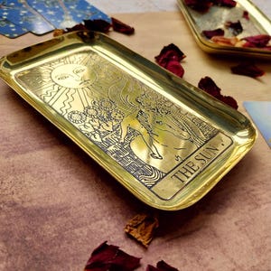 Pu&ograve; includere: Un vassoio rettangolare in ottone dorato con angoli arrotondati, con un disegno inciso del sole, una figura e la scritta "THE SUN". Petali di rosa secchi e carte dei tarocchi sparsi intorno.
