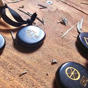 Op de afbeelding: Vijf gladde zwarte stenen met gouden symbolen en tekst. De stenen zijn gerangschikt op een houten oppervlak. De symbolen omvatten een driehoek, een cirkel met een lijn erdoorheen en een vierpuntige ster. De tekst op de stenen is "AIR", "FIRE", "EARTH", "WATER" en "SPIRIT".