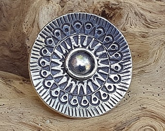 May include: A silver-toned metal ring with a circular design featuring a central circle and concentric circles of intricate patterns.