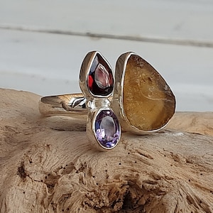 Könnte beinhalten: Ein silberner Ring mit drei Edelsteinen: einem violetten Amethyst, einem roten Granat und einem gelben Citrin. Der Ring liegt auf einem Stück Treibholz.