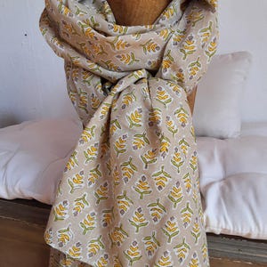 Peut inclure: Écharpe beige avec un motif répété de formes en forme de feuilles jaunes et vertes. L'écharpe est drapée sur une forme en bois, mettant en valeur sa texture douce et son design. La couleur neutre de l'écharpe la rend polyvalente.