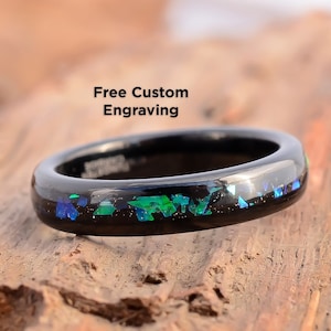 Könnte beinhalten: Schwarzer Ring mit einem Band aus schimmerndem grünem und blauem Glitzer, eingebettet in schwarzem Harz. Der Ring liegt auf einer Holzoberfläche. Kostenlose Gravur nach Wunsch.