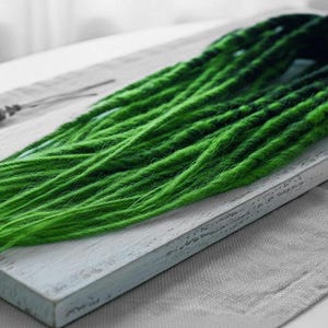 May include: Bright green dreadlocks, arranged on a white wooden board. The dreadlocks are a vibrant green, with darker green at the roots, creating a gradient effect. The board rests on a white linen surface, with decorative elements.