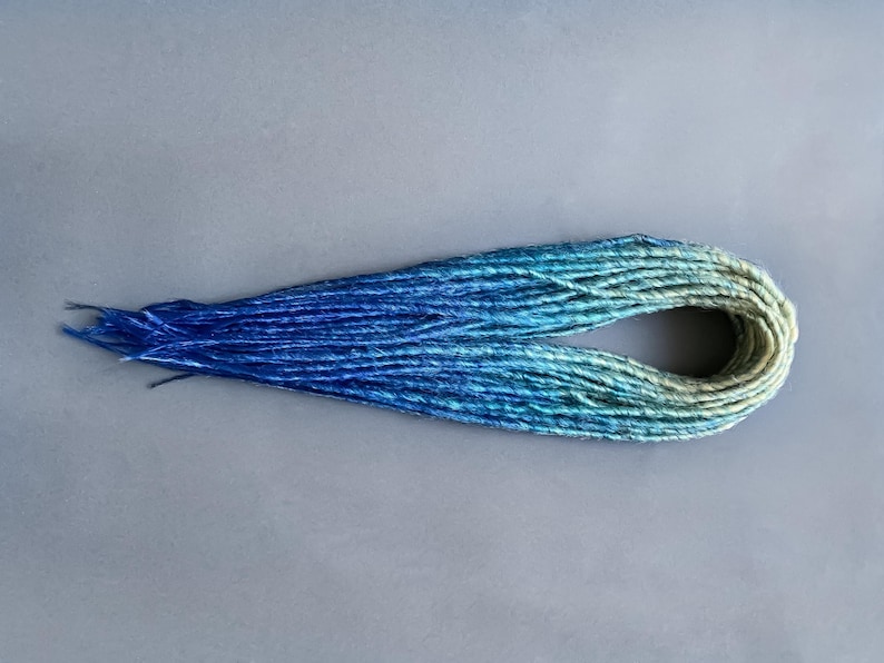 May include: A set of ombre synthetic dreadlocks in shades of blue and green. The dreadlocks are arranged in a loose circle.