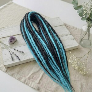 May include: A bundle of black and turquoise dreadlocks. The dreadlocks are arranged on a white wooden surface, with a light-colored fabric underneath. A small purple crystal and sprigs of lavender and white flowers are also present.