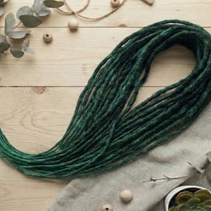 Puede incluir: Rastas de color verde oscuro, dispuestas en un haz suelto, sobre una superficie de madera clara. Las rastas tienen una apariencia texturizada y un degradado de tonos verdes. Ramas de eucalipto y pequeñas plantas suculentas.