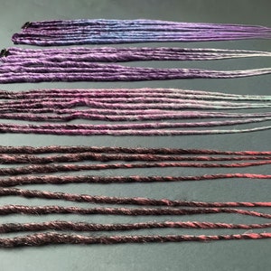 May include: A set of synthetic dreadlocks in various shades of purple, pink, and black. The dreadlocks are arranged in rows, with the purple and pink dreadlocks at the top and the black dreadlocks at the bottom.