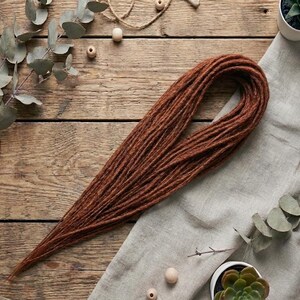 May include: A single, long, brown dreadlock, resting on a light gray cloth and a wooden surface. The dreadlock is a warm, reddish-brown color. The background includes eucalyptus branches, small succulents in white pots, and wooden beads.