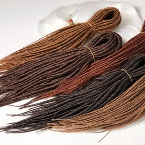 Puede incluir: Cuatro paquetes de rastas sintéticas en varios tonos de marrón, incluyendo marrón oscuro, marrón claro y marrón rojizo. Las rastas están dispuestas en forma de abanico sobre una superficie blanca.