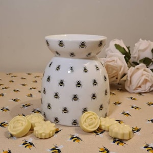 May include: White ceramic wax melt warmer with a bee pattern. The warmer has a round base and a wide, flat top. There are six white wax melts on a beige surface with a bee pattern.