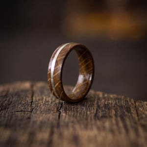A ring crafted from brown oak wood featuring a smooth, polished silver inlay that runs horizontally around the center of the band. The wood grain is visible, showing a natural texture and deep brown tones throughout the ring.