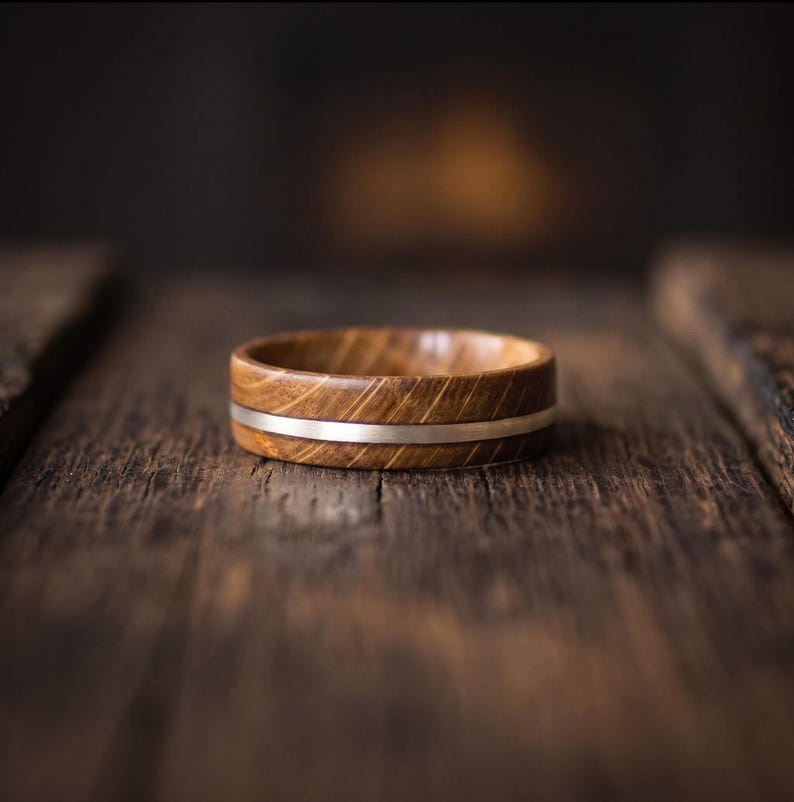 A ring crafted from brown oak wood with a visible grain pattern, featuring a thin, smooth, horizontal silver metal band inlaid around the center of the exterior circumference of the circular wooden ring body.
